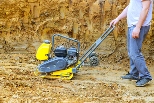 Construction worker pushes plate compactor over loose soil at building site in dust rises as machine flattens ground for paving.