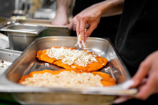 Close-up of a person's hand sprinkling shredded cheese onto traditional Hungarian langos in a professional kitchen setting. Fried dough street food preparation