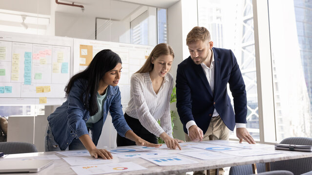 Three multiethnic teammates, marketing analysts gathered around conference desk in office, analyzing charts and graphs, reviewing campaign metrics, performance data in documents spread across table