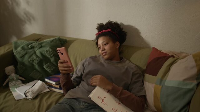 Handheld shot of dreamy African American teenage girl lying on couch holding notebook surrounded by educational materials and using smartphone, checking messages while studying at home