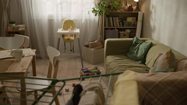 Handheld no people full shot of modest family apartment interior featuring baby feeding chair, laundry drying on rack, small dining table and childrens drawings on fridge