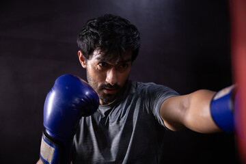 Determined male boxer training hard punching heavy red bag wearing blue boxing glove looking...