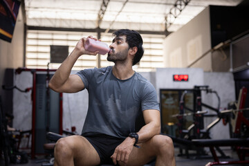 Obraz premium Tired young man drink water from bottle to rest after heavy workout in gym. Athlete covered in sweat look exhausted but stay determined to achieve strong fitness health goal