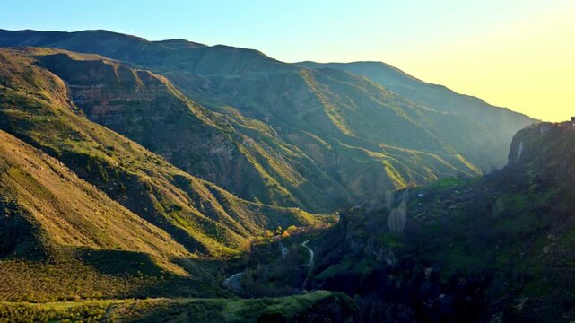 Sunlight bathes rolling mountain slopes in warm hues. Greenery clings to rugged terrain under golden rays. Valley winds drift through deep gullies below