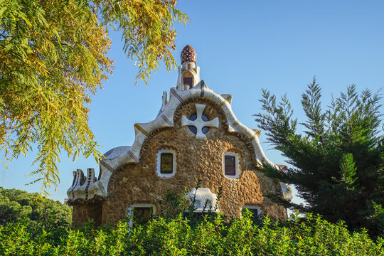 Park Guell,Barcelona,Spain,A unique stone building with organic architectural design, featuring a cross emblem and surrounded by greenery under a clear blue sky.