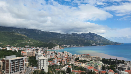 Fototapeta premium Mountains overlook a coastal town with buildings and water on a cloudy day. Budva, Montenegro.