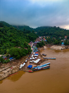Aerial view of longtail boats and white tents along the muddy riverbank of the Mekong River surrounded by misty green hills in Pak Beng, Oudomxay Province, Laos.