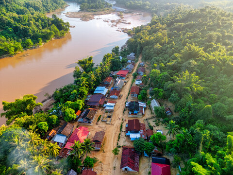 Aerial view of a small village with colorful rooftops nestled along a winding river surrounded by dense tropical forest in Nong Khiaw, Luang Prabang Province, Laos.