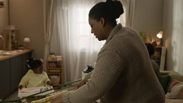 Arc shot of caring Black mother hanging laundry on drying rack in small modest apartment with teenage girl entertaining her little sister sitting in high chair