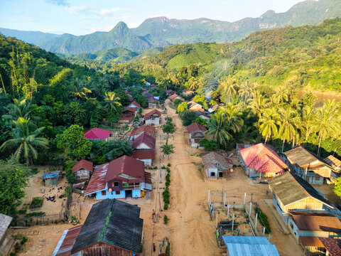 Aerial view of a small village with traditional houses and a dirt road nestled among lush tropical forests and limestone mountains in Nong Khiaw, Luang Prabang Province, Laos.