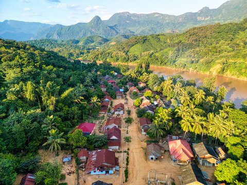 Aerial view of a small village with red-roofed houses nestled between lush green forests and the Nam Ou river under limestone karst mountains in Nong Khiaw, Luang Prabang Province, Laos.
