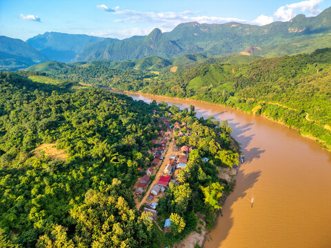 Aerial view of a small village along the Nam Ou River surrounded by dense tropical forest and limestone mountains in Nong Khiaw, Luang Prabang Province, Laos.