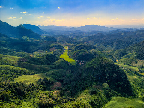 Aerial view of lush green mountains and a valley with vibrant rice paddies under a blue sky in Nong Khiaw, Luang Prabang Province, Laos.