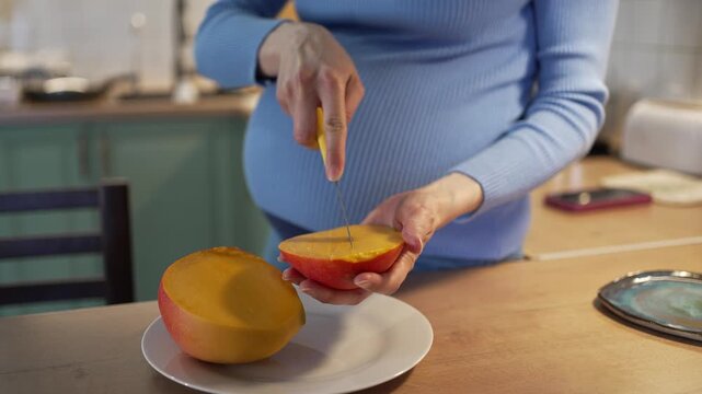 Pregnant woman carefully slicing a fresh, ripe mango in her kitchen, highlighting the importance of a nutritious diet, vitamins, and healthy eating habits during maternity for her baby's growth