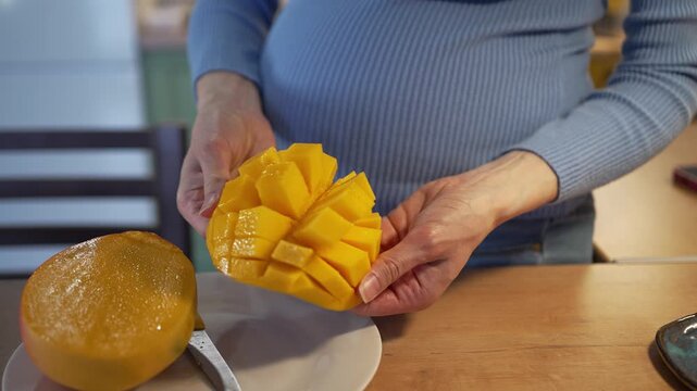 Pregnant woman carefully cutting a fresh, ripe mango into cubes with a knife, highlighting a healthy diet and proper nutrition essential for maternal wellbeing during her expectancy