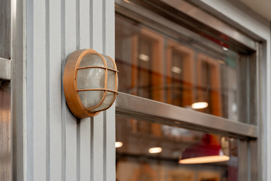 Industrial bulkhead wall light with a metal cage mounted on a grooved gray facade next to a modern metal-framed window.