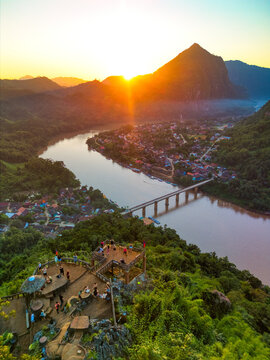 Aerial view of the Nam Ou River winding through the village and mountains with people at a viewpoint during sunset in Nong Khiaw, Luang Prabang Province, Laos.