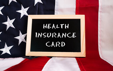 Fototapeta premium Health Insurance Card chalkboard sign on American flag background. USA healthcare insurance policy reform concept