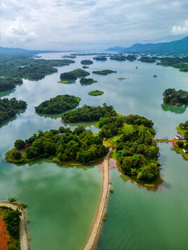 Aerial view of the Nam Ngum Reservoir featuring lush green islands scattered across calm waters with a narrow dirt road and distant mountains under a cloudy sky in Laos.