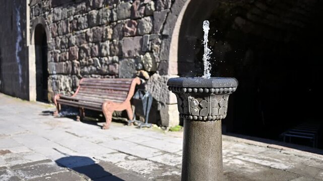 Water fountain sprays gently on stone pedestal. Wooden bench sits beside ancient stone wall. Sunlight casts shadows on worn pavement. Arched doorway leads into dark tunnel. Scene feels peaceful