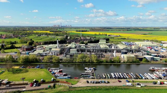 Aerial view of industrial chemical production factory on river bank with narrowboats docked and power station towers in the background and sunny rapeseed fields UK