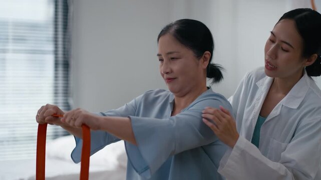 Caring young Asian women doctor smiling and encouraging elderly female patient doing arm stretching exercise with resistance band in clinic.Concept of physical therapy,rehabilitation,Physical Medicine