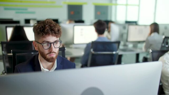 Young businessman in glasses working at desktop computer in modern open space office. Focused male office employee analyzing information on screen while colleagues work in the background. Professional