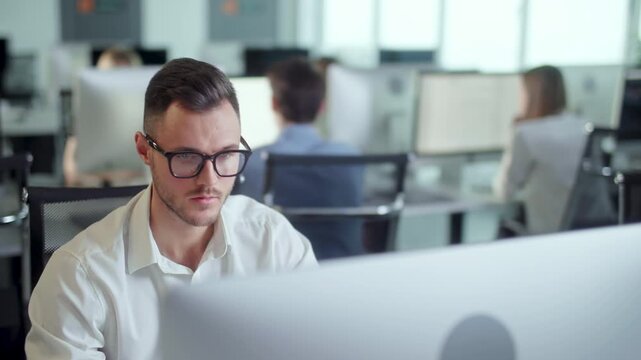 Serious young office employee in glasses working at computer in modern open space office. Concentrated businessman looks at monitor while colleagues work in the background. Professional corporate