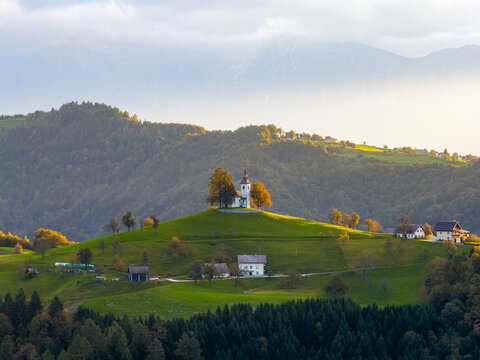Aerial view of St. Thomas's Church on a green hilltop surrounded by autumn trees and misty mountains in Ratece Planica, Jesenice, Slovenia.