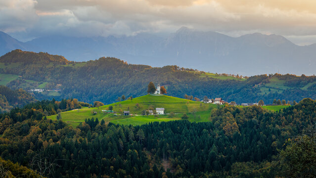Aerial view of St. Thomas's Church on a lush green hilltop surrounded by dense forests and the Julian Alps under a cloudy sky in Ratece Planica, Jesenice, Slovenia.