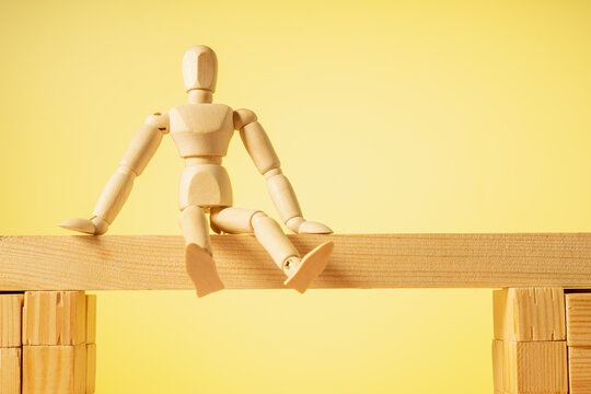 A wooden figure sits on a yellow background with its legs hanging down