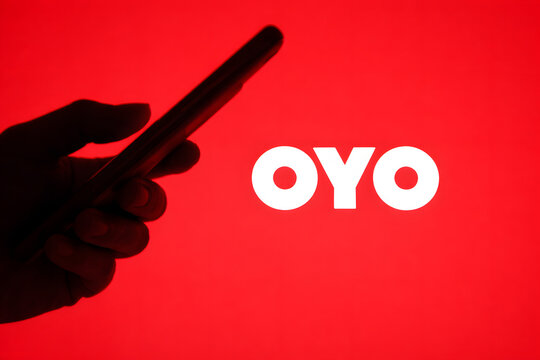 OYO Hotels brand logo illuminated on red background with silhouette of hand holding smartphone, Indian hospitality startup and travel technology company concept
