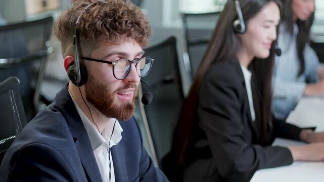 Young male customer support agent speaking through headset while working at computer in a modern call center office. Professional help desk employee communicates with clients, while colleagues in the