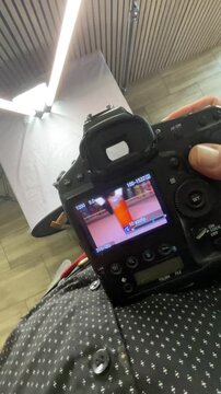 Vertical stock video camera display showing food previews on DSLR screen with photographer wearing patterned shirt, studio umbrella and backdrop in frame, hands adjusting settings,