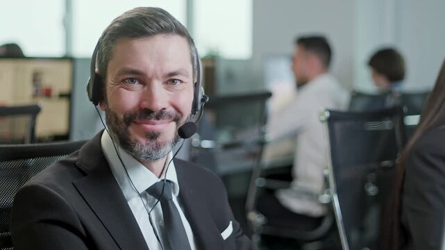 Smiling mature male customer support agent wearing headset and looking at camera while working in a modern call center office. Friendly professional operator in formal business suit represents