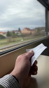 Passenger holds ticket by window. Person with ticket looking outside at passing village. Traveling individual holding ticket gazing at passing village houses with soft light