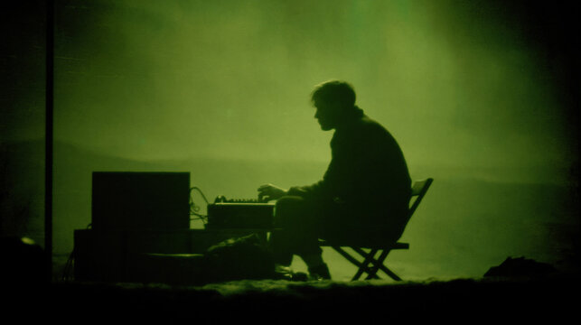 Solitary Contemplation: In a scene veiled in an emerald hue, a figure finds solace in solitude, engrossed in tasks by a cryptic electronic device amidst an atmospheric backdrop. 