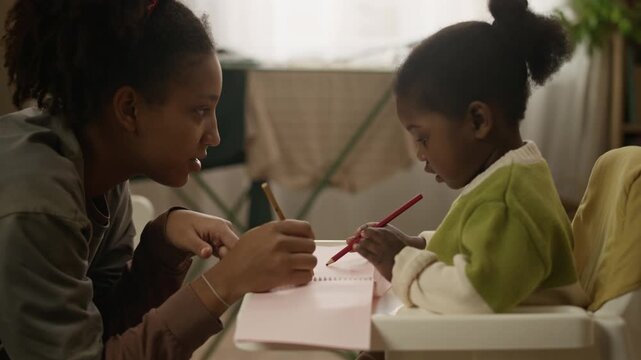 Handheld side view shot of African American teenage girl drawing in notebook with playful toddler girl sitting in high chair, taking care of little sister in modest small family apartment