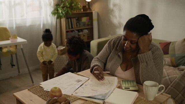 Handheld shot of young Black mother sitting at table at home and calculating monthly expenses while reviewing overdue utility bills, with her teenage daughter playing with toddler girl in background
