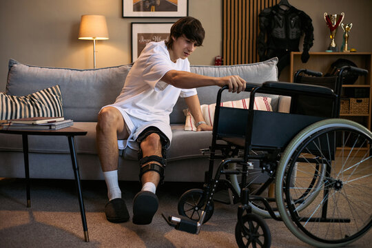Young adult Caucasian man with disability reaching for wheelchair while recovering at home with leg brace, representing mobility support and rehabilitation. Useful for healthcare campaigns