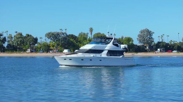 White luxury motor yacht cruises through calm blue bay waters on clear sunny day. Palm trees and green vegetation line sandy shoreline with parked vehicles visible.