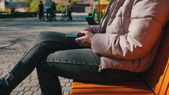 An unrecognizable woman sits cross-legged on an orange wooden bench and uses her smartphone. She wears a casual puffer jacket on a bright sunny day. Blurred cyclists ride by in the background.