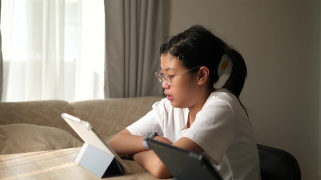 Focused young Asian girl wearing glasses using a digital tablet and stylus for online learning at home with warm sunlight background, education and technology concept