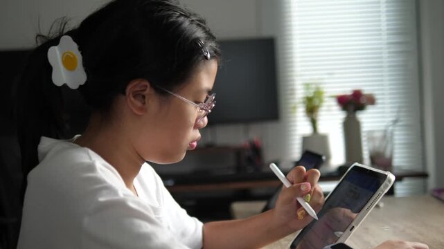 Profile view of a focused young Asian girl wearing glasses and a cute hair clip, using a digital tablet and stylus for online learning at home