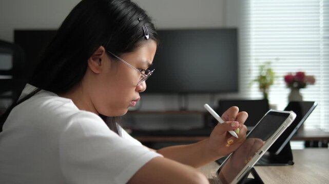 Profile view of a focused young Asian girl wearing glasses using a digital tablet and stylus for online learning at a desk, modern education concept
