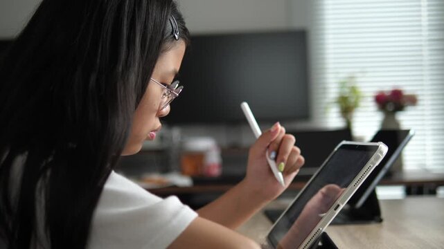 Profile view of a focused young Asian girl wearing glasses using a digital tablet and stylus for online learning at a desk, modern education concept
