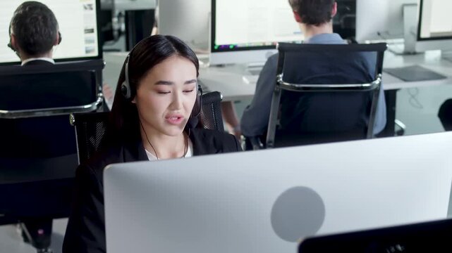 Young female customer support agent wearing headset and working at computer in a modern open plan call center office. Professional businesswoman handles client communication and support requests while
