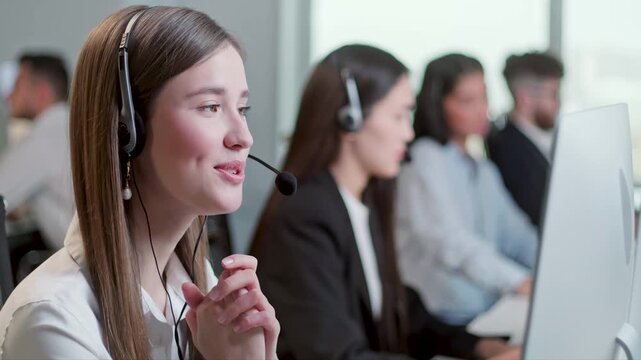 Young female customer support agent wearing headset and smiling while speaking with client in a modern call center office. Professional support operator gestures with hands during conversation