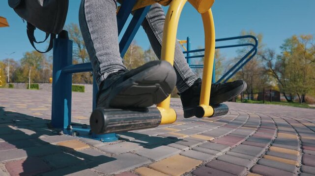 Legs of a person in sneakers pushing pedals on an outdoor yellow exercise machine in a sunny public park. Represents an active lifestyle and community health concept.