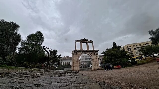 Wide shot of the ancient monumental Arch of Hadrian in Athens, Greece, during a cloudy and rainy day, with tourists carrying umbrellas walking through the archaeological site.